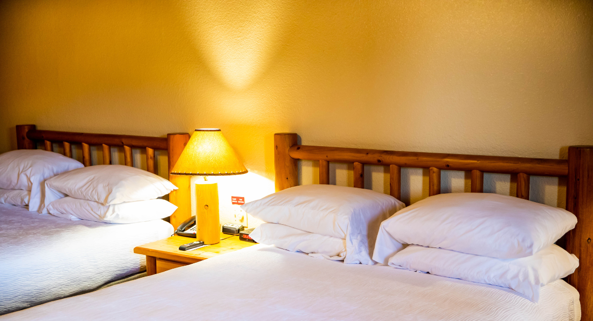 A hotel room with two beds, a wooden frame, white bedding, and a lamp on a nightstand.