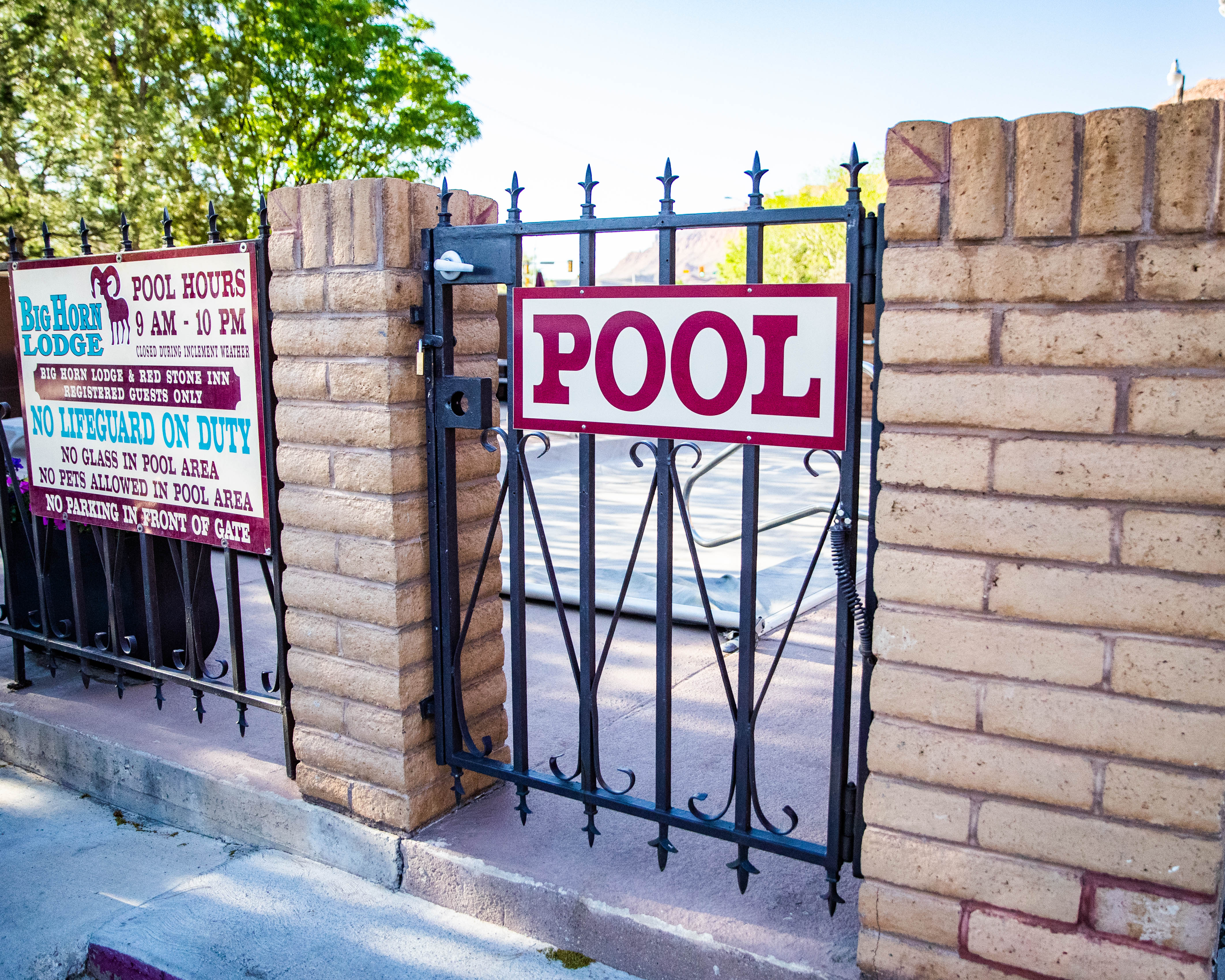A metal gate with a red and white sign reading 'POOL' and a brick wall with another sign listing pool rules and hours.